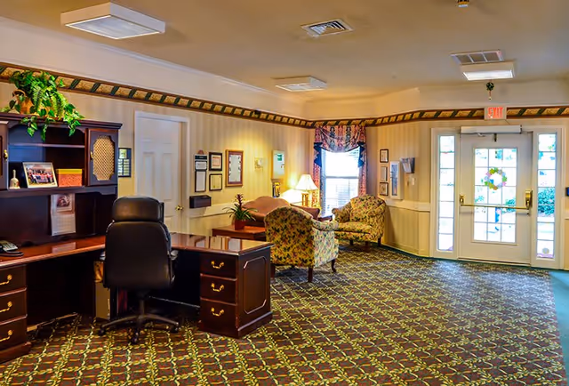 A well-lit interior room with patterned carpet, a dark wooden desk with a black office chair, two floral upholstered armchairs, a small table with a lamp, framed pictures on the walls, and double glass doors with decorative decals leading outside.