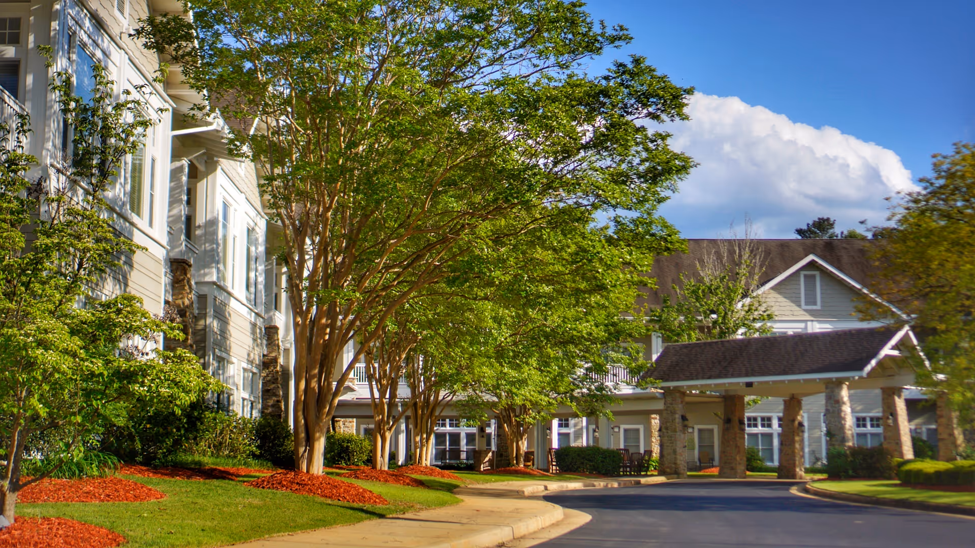 Exterior view of Vernon Woods senior living facility showing a driveway lined with green trees and landscaped areas with red mulch. The building has light-colored siding, multiple windows, and a covered entrance supported by stone pillars under a clear blue sky with some clouds.
