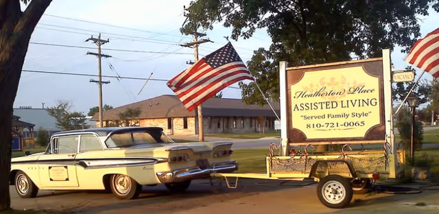 A vintage yellow car is parked next to a trailer displaying a sign for Heatherton Place Assisted Living with the tagline 'Served Family Style' and a phone number. Two American flags are mounted on the trailer and nearby. There is a building and trees in the background under a clear sky.