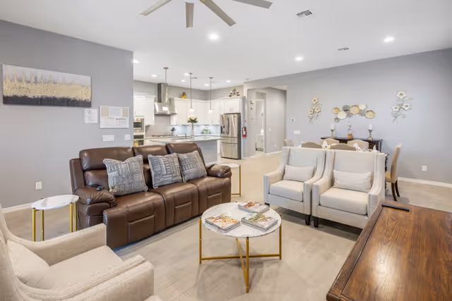 A bright and spacious living room area with a brown leather sofa, two beige armchairs, and a round marble coffee table with magazines on it. The room has light-colored wood flooring and gray walls decorated with artwork. In the background, there is an open kitchen with white cabinets, stainless steel appliances, and pendant lighting. A dining area with a table and chairs is also visible.