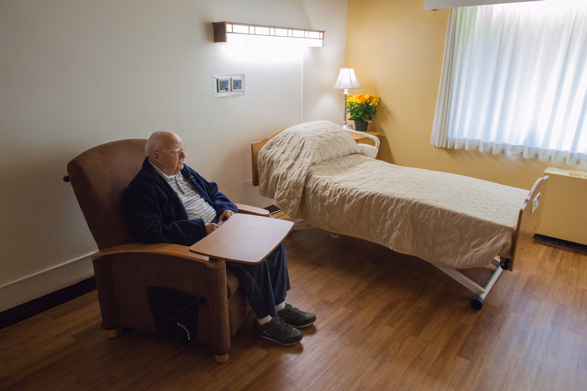 An elderly man sitting in a brown recliner chair with a small attached table in a room with a single bed covered with a beige quilt. The room has wooden flooring, a small bedside table with a lamp and a potted plant with yellow flowers, and a window with white curtains letting in natural light.