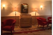 Cozy lounge with red upholstered chairs, a wooden bureau between two floor lamps, a framed painting, and wall sconces over a patterned rug.