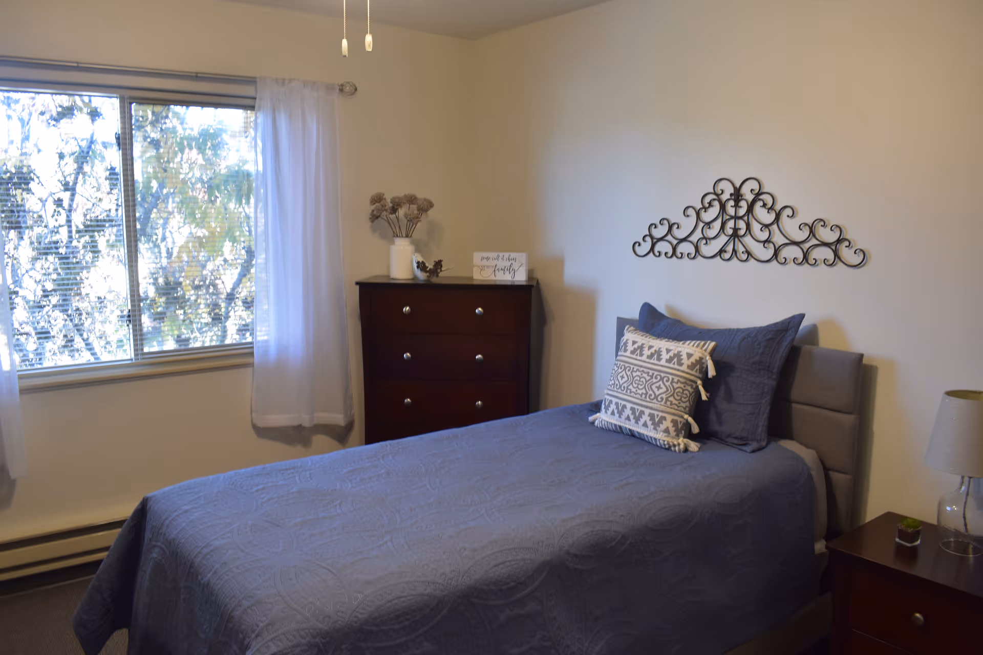 A bedroom with a single bed covered in a blue quilt and two decorative pillows. There is a dark wooden dresser with a vase of dried flowers and a small sign on top. A window with white curtains lets in natural light, and a decorative metal wall art piece hangs above the bed. A bedside table with a lamp and a small plant is next to the bed.