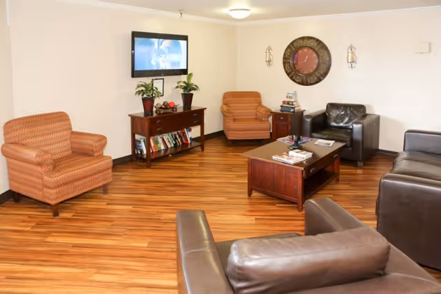 A cozy living room with wooden flooring, featuring two patterned armchairs, three dark leather armchairs, a wooden coffee table with magazines, a wooden console table with books and plants, a wall-mounted TV, a large decorative clock, and two wall sconces.