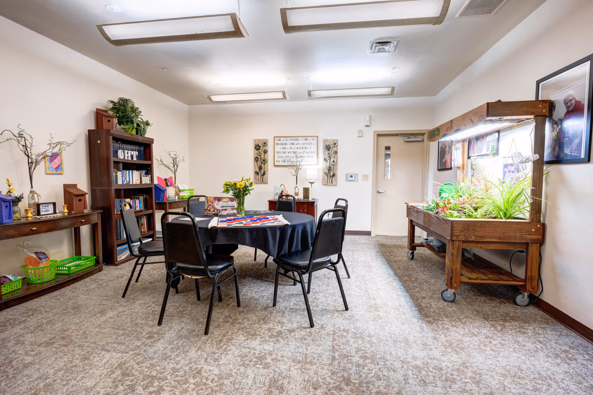 A well-lit room with a round table covered by a black tablecloth and surrounded by five black chairs. The table has a vase with yellow flowers and a board game on it. To the right, there is a wooden plant stand with various green plants under grow lights. On the left side, there is a wooden bookshelf filled with books and decorative items, and a long wooden table with green baskets and small decorations. The walls are light-colored with framed pictures and wall art. The floor is carpeted in a neutral pattern.