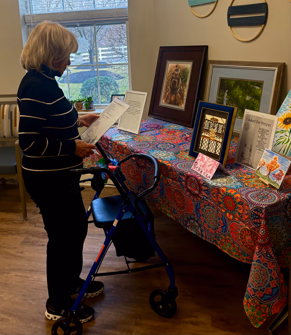 An elderly woman with blonde hair using a blue walker is standing indoors near a table covered with a colorful patterned cloth. On the table are framed pictures, artwork, and printed poems or writings. A window with blinds and a view of bushes and a white fence is in the background.