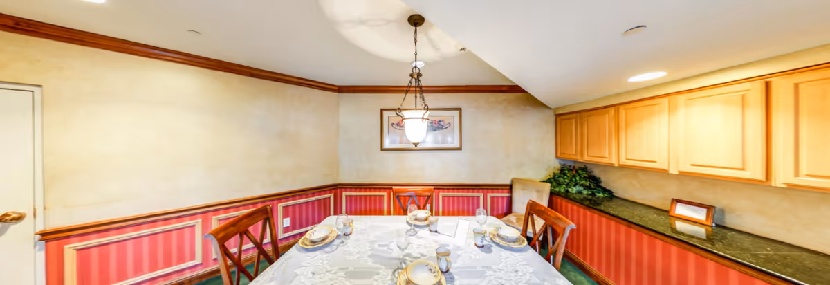 A dining room with a rectangular table set for four people with plates, glasses, and napkins. The room features red and beige striped wallpaper with wooden trim, a hanging light fixture above the table, wooden chairs, and a long countertop with cabinets above it on the right side. There is a door on the left wall and a framed picture hanging on the back wall.