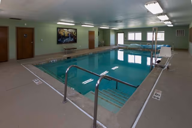 Indoor swimming pool in a green-walled room with metal handrails, a pool lift, benches and fluorescent ceiling lights.