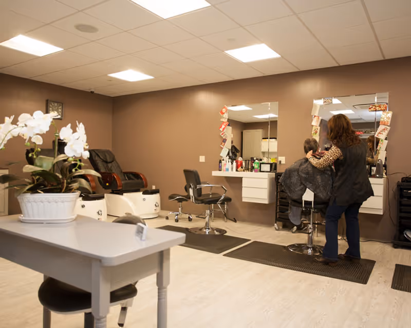 Interior view of a salon area in a senior living facility with a hairdresser cutting a person's hair in front of a large mirror. The room has brown walls, white ceiling tiles with recessed lighting, and light wood flooring. There are salon chairs, a pedicure station, and a table with a white orchid plant in the foreground.