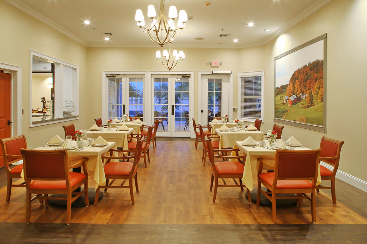 A dining room with wooden floors and cream-colored walls, featuring six tables covered with beige tablecloths, each set with napkins, glasses, and small flower arrangements. The chairs have wooden frames with red cushions. Two chandeliers hang from the ceiling, and large windows and glass doors at the far end let in natural light. A large framed picture of a rural landscape with trees and barns is on the right wall.