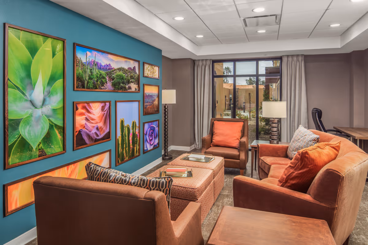 A cozy living room area with brown armchairs and a sofa adorned with orange and patterned cushions. A pair of ottomans serve as a coffee table with books and trays on top. The walls are decorated with colorful framed nature photographs, and a large window with curtains offers a view of an outdoor courtyard. The room has a modern ceiling with recessed lighting.