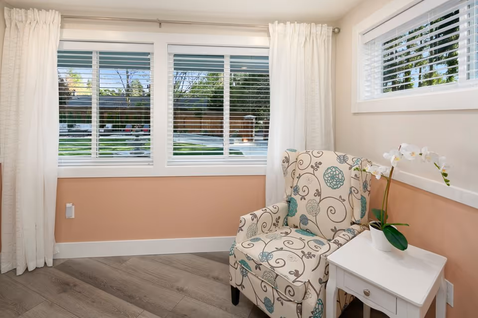A cozy corner in a senior living facility featuring a patterned armchair with floral designs next to a small white side table holding a white orchid plant. The room has light peach walls with white trim, large windows with white blinds and curtains, and a view of a green outdoor area.