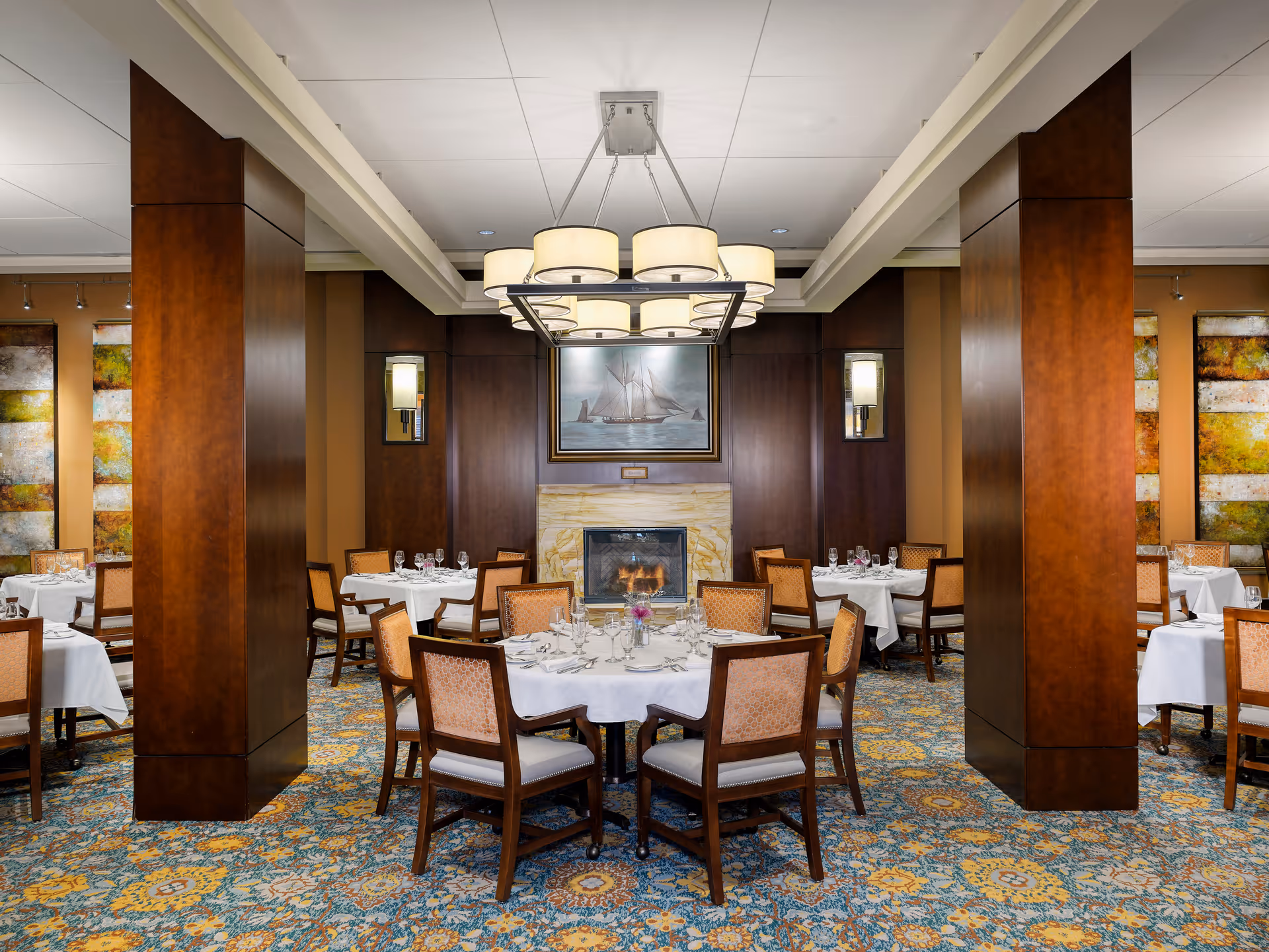 Elegant dining room with round tables covered in white tablecloths, set with glassware and silverware. The room features wooden pillars, a patterned carpet, a fireplace with a painting of a sailboat above it, and a modern chandelier hanging from the ceiling.
