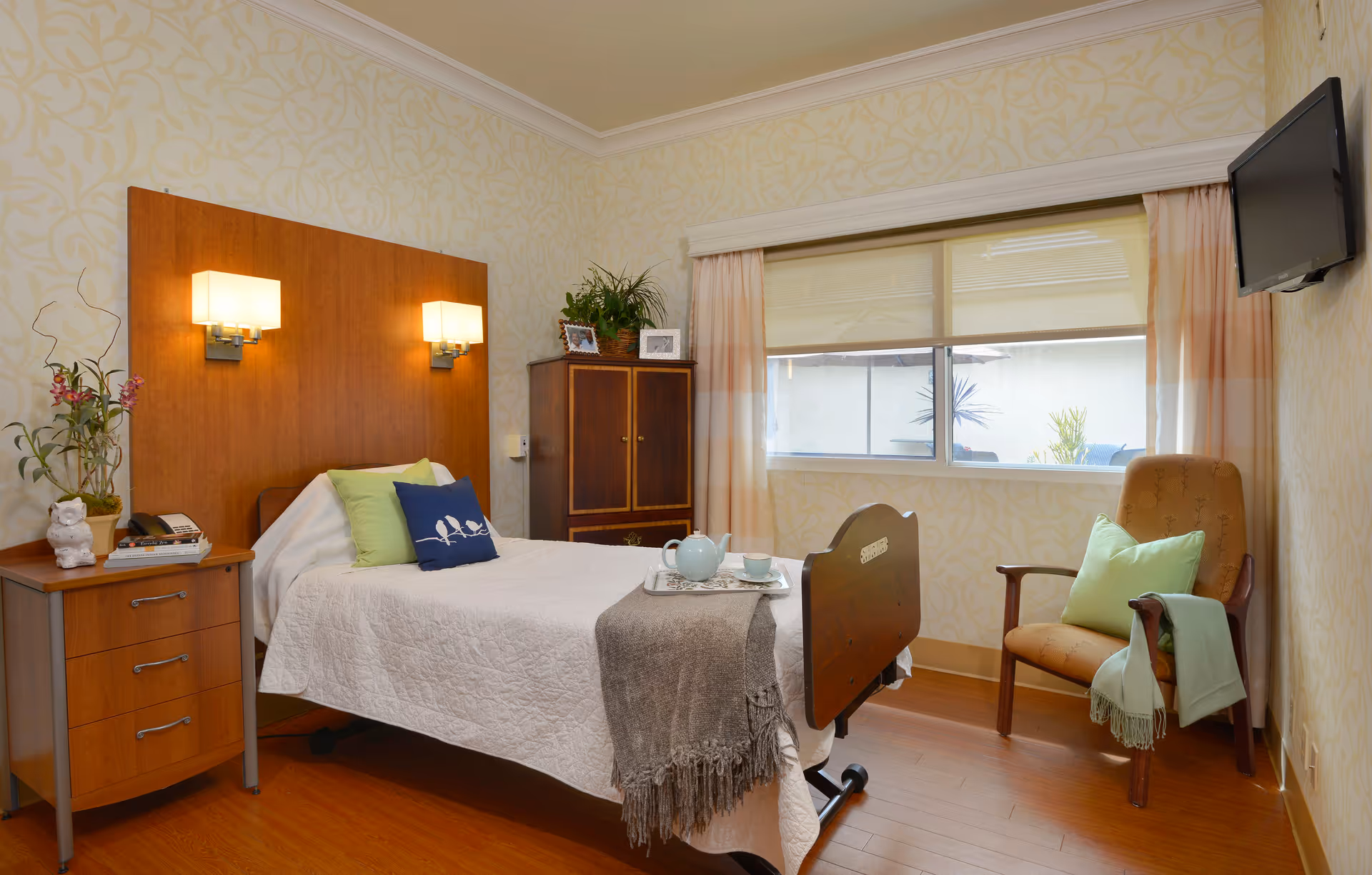 A cozy bedroom in a senior living facility featuring a single bed with white bedding, green and blue pillows, and a gray throw blanket. The room has wooden flooring, a wooden headboard with two wall-mounted lamps, a wooden nightstand with a plant and books, a wooden cabinet with framed photos and a plant on top, a window with curtains, a comfortable armchair with a green pillow and a folded blanket, and a wall-mounted TV.