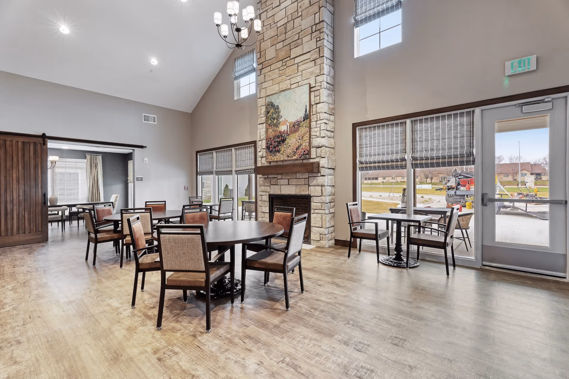 Bright communal dining room with round tables and chairs, large windows, and a tall stone fireplace topped with artwork.