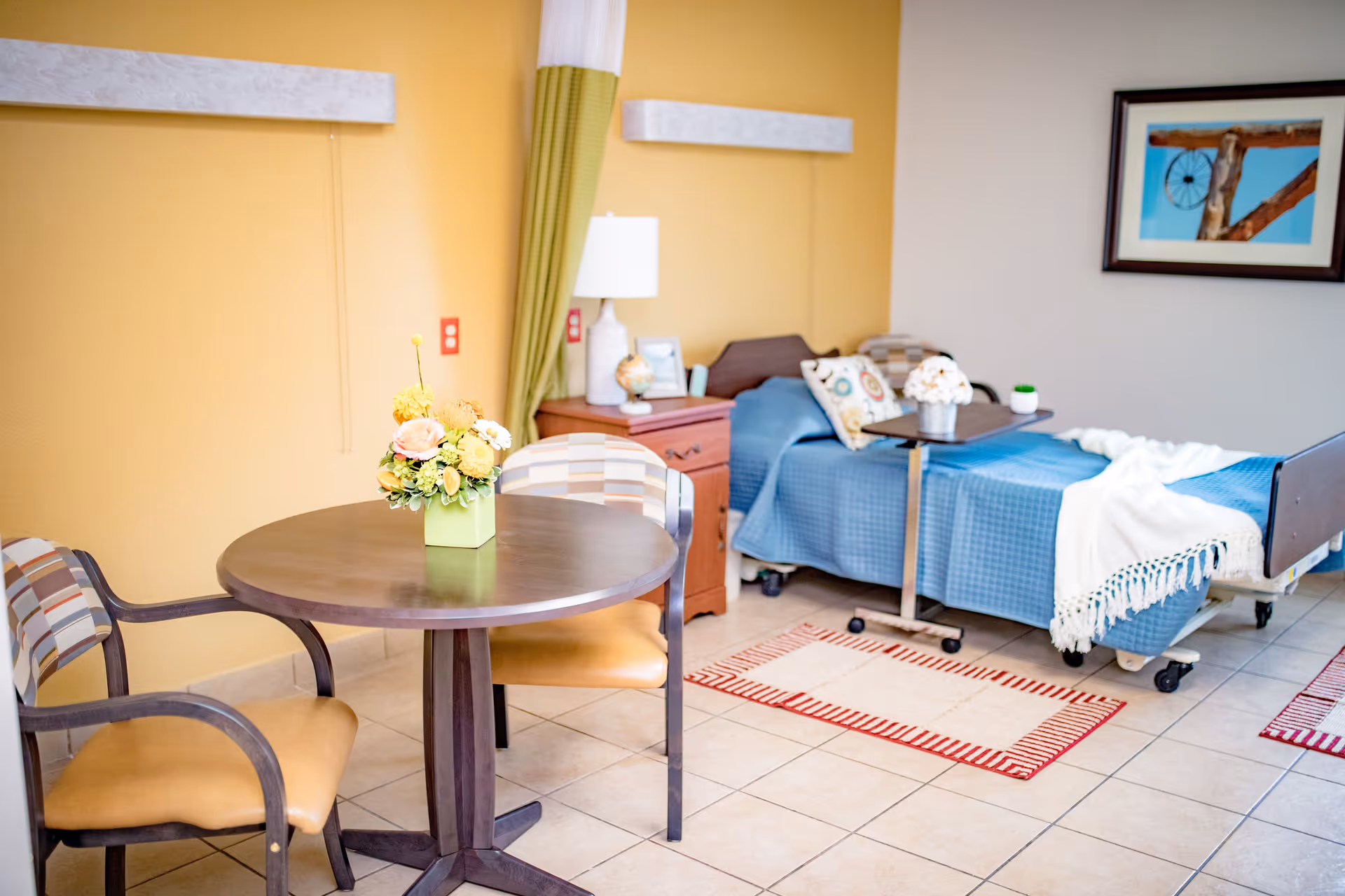 A bright and cozy nursing facility room with a single bed covered in a blue blanket and white throw, a wooden nightstand with a lamp and decorative items, a small round table with a flower arrangement, and two chairs with patterned cushions. The walls are painted yellow and beige, and a framed picture hangs on the wall.