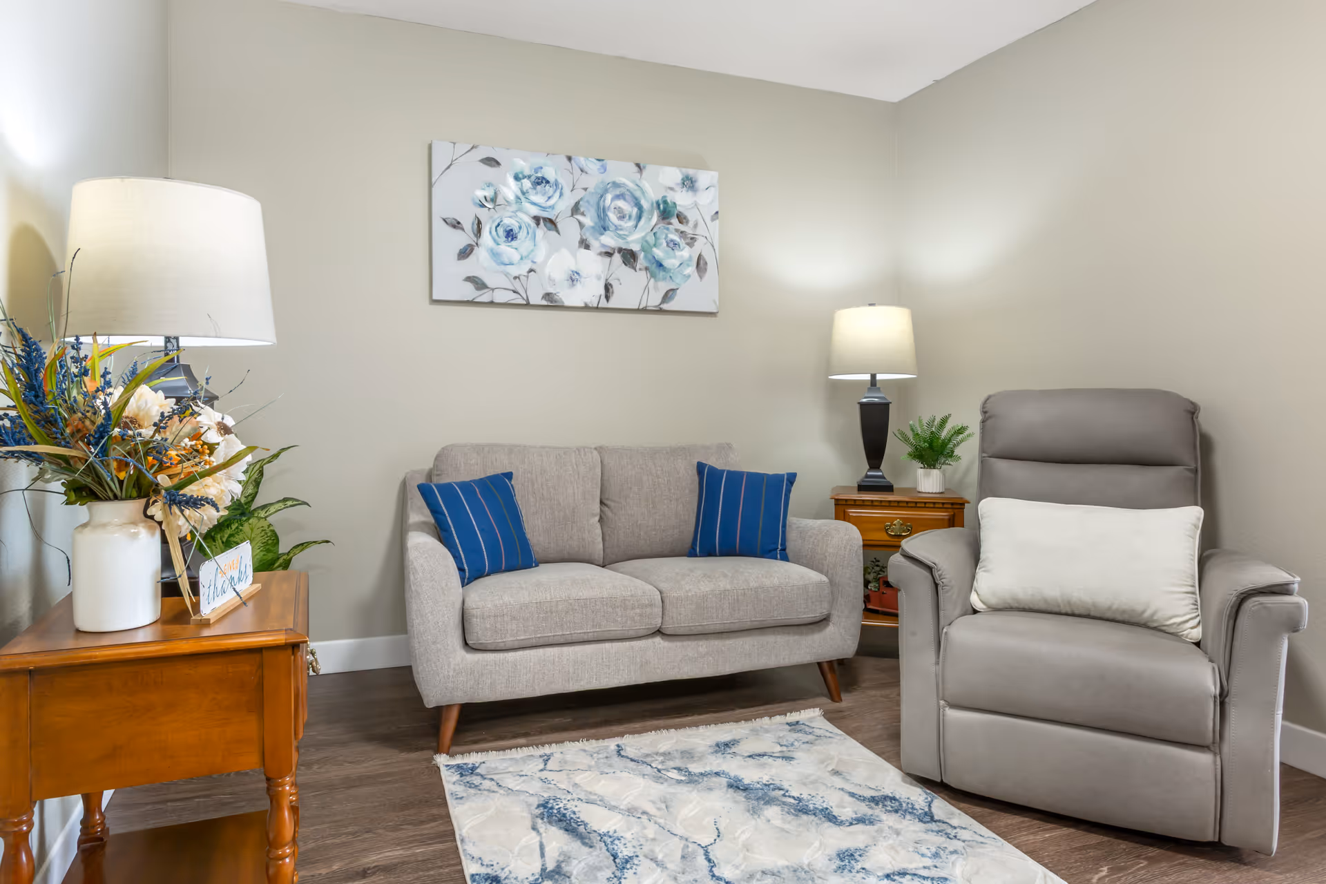 A cozy living room area featuring a light gray loveseat with two blue striped pillows, a gray recliner chair with a white pillow, a wooden side table with a lamp and a floral arrangement, another wooden side table with a lamp and a small potted plant, and a decorative rug with blue and white patterns on a wooden floor. A floral painting with blue flowers hangs on the beige wall behind the loveseat.