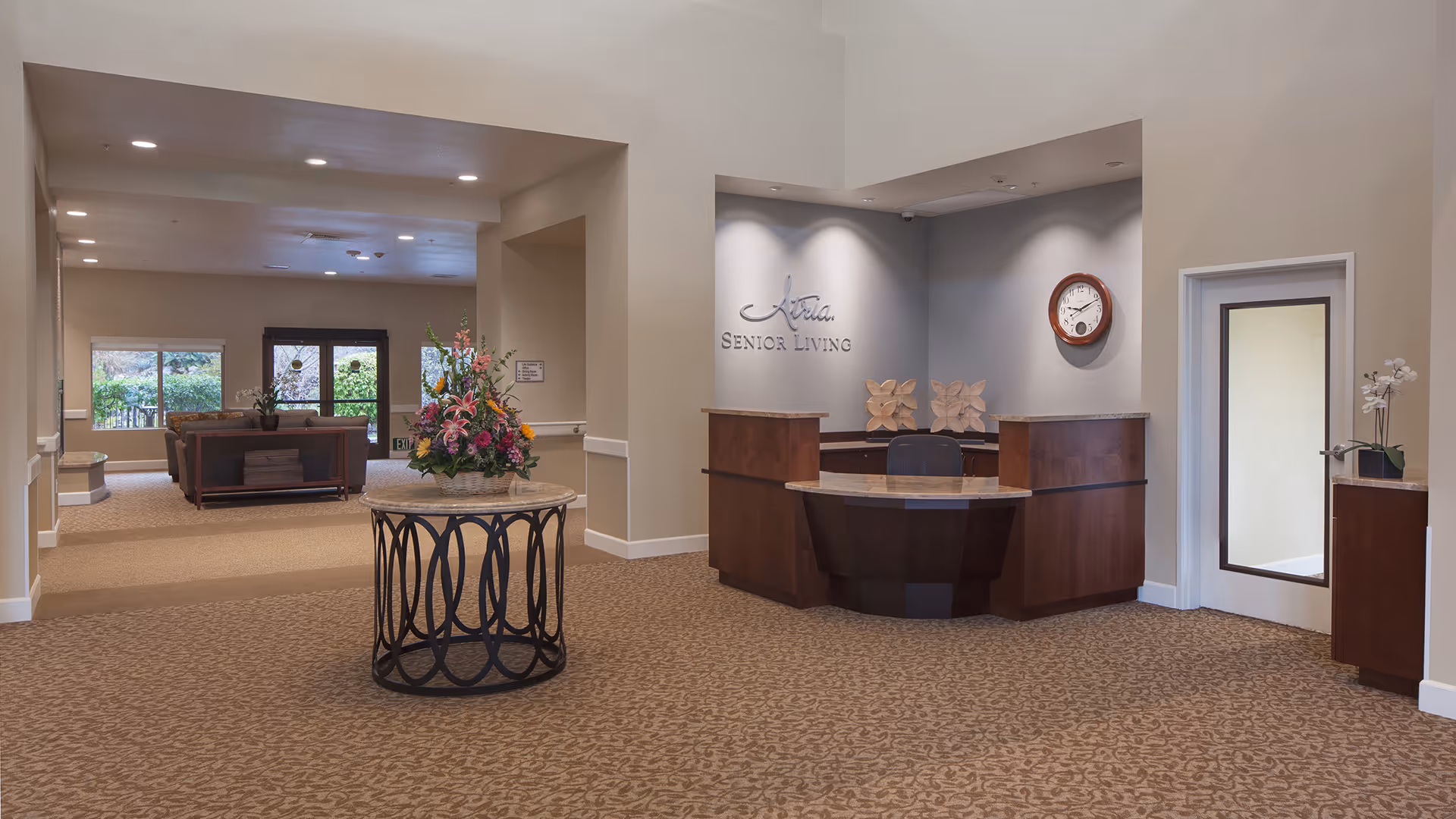 Reception area of Atria Walnut Creek senior living facility with a wooden front desk, a round table with a floral arrangement in the center, beige walls, carpeted floor, and a seating area with sofas and large windows in the background.