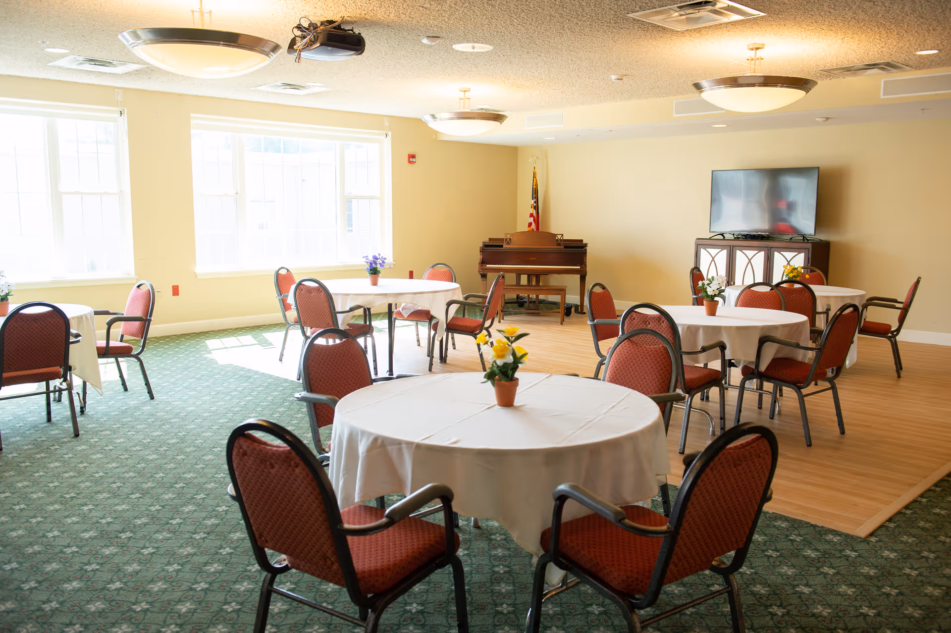 A bright room with round tables covered in white tablecloths, each surrounded by red cushioned chairs. Small flower pots decorate the tables. The room has large windows letting in natural light, a green carpeted floor with a wooden section, a piano against the far wall, and a flat-screen TV on a cabinet.