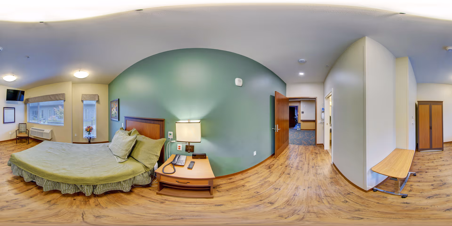 A spacious bedroom in a senior living facility with a large bed covered in green bedding, a wooden nightstand with a lamp, telephone, and remote control. The room has wooden flooring, a green accent wall behind the bed, a window with blinds and a valance, a small table with a flower vase, a wall-mounted TV, and an open door leading to a hallway.