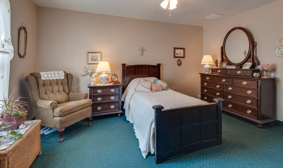 A cozy bedroom with a single wooden bed covered with a white quilt and decorative pillows. To the left of the bed is a wooden nightstand with a floral lamp and an alarm clock. Next to the nightstand is a cushioned armchair with a doily on the backrest. On the right side of the bed is a large wooden dresser with an oval mirror, a lamp, and decorative items. The room has green carpet flooring and beige walls with framed pictures and a small cross hanging above the bed.