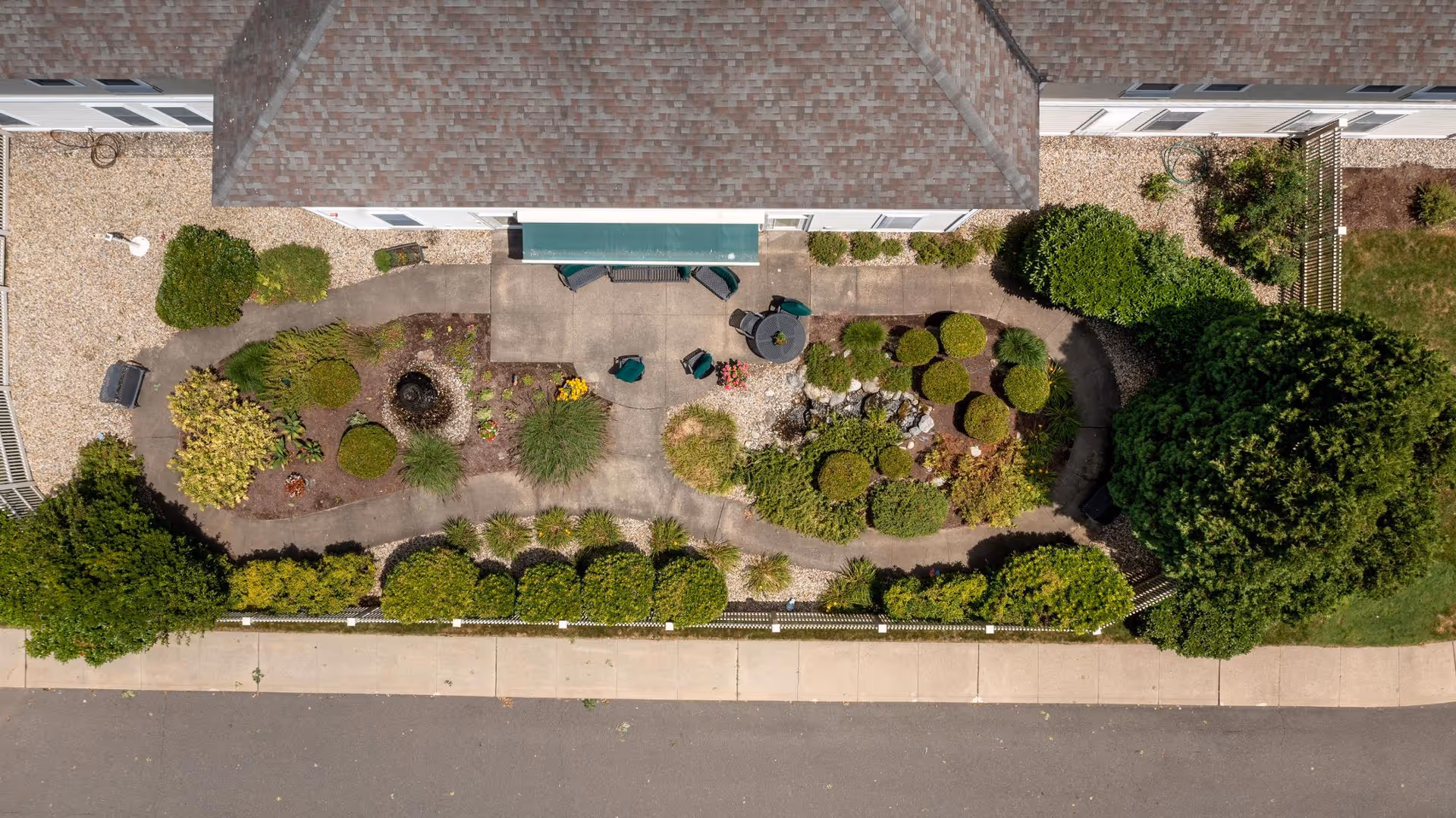 Aerial view of a landscaped garden area behind a building with a paved walkway, various bushes, shrubs, and a small seating area with a table and chairs. The garden is bordered by a sidewalk and a road.