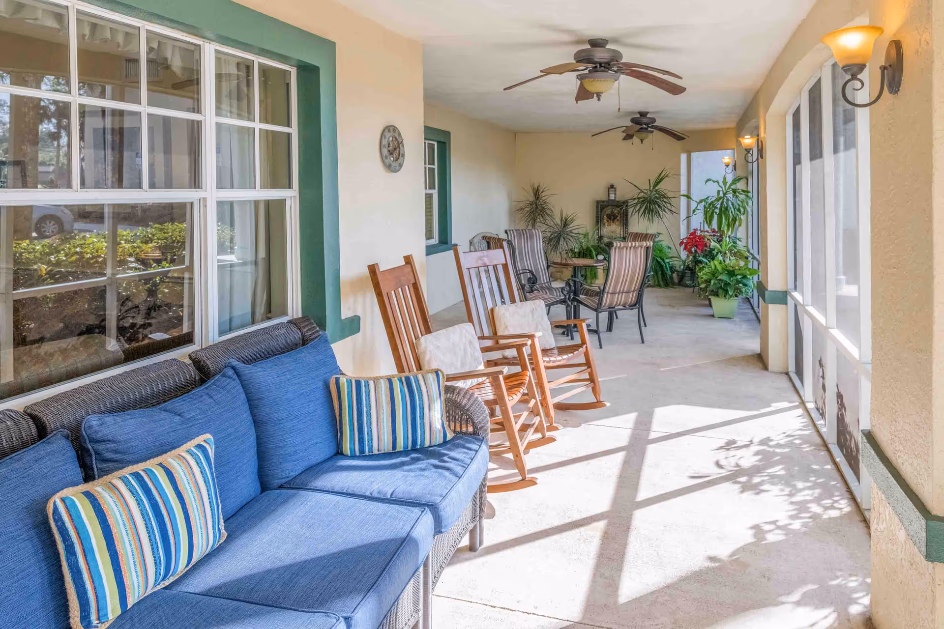 A covered outdoor patio area with a blue cushioned wicker sofa, two wooden rocking chairs with cushions, and a round table with four striped cushioned chairs. The patio is decorated with several potted plants and has ceiling fans and wall-mounted lights.