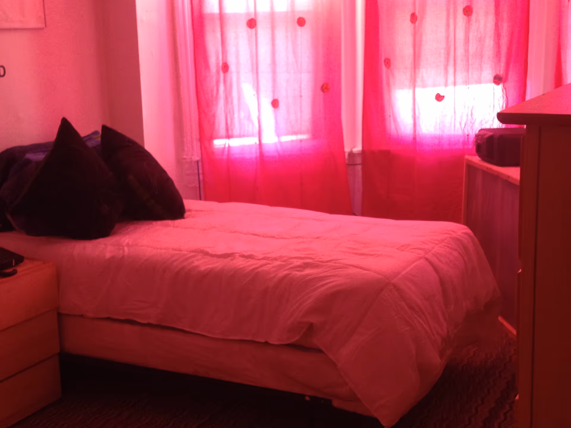 A bedroom with a single bed covered in a white comforter and two dark pillows. The room has red curtains with circular patterns covering the window, a wooden dresser on the right side, and a small nightstand on the left side.