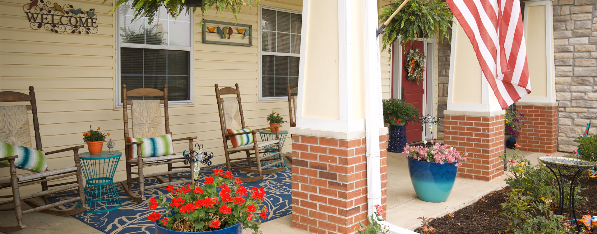 Front porch area of a senior living facility with three wooden rocking chairs, each with a colorful cushion, placed on a blue patterned rug. There are potted plants with red and orange flowers on small tables and on the ground. The porch has cream-colored siding, brick pillars, a red door decorated with a wreath, and an American flag hanging from one of the pillars. A 'WELCOME' sign and a decorative bird wall art are mounted on the wall above the chairs.