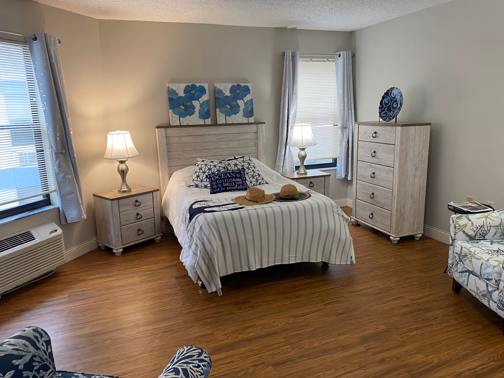 A cozy bedroom with a wooden bed frame, white and blue bedding, two straw hats on the bed, two bedside tables each with a lamp, a wooden dresser with a decorative plate on top, two windows with light curtains, and a patterned armchair in the corner. The floor is wooden and the walls are light-colored.
