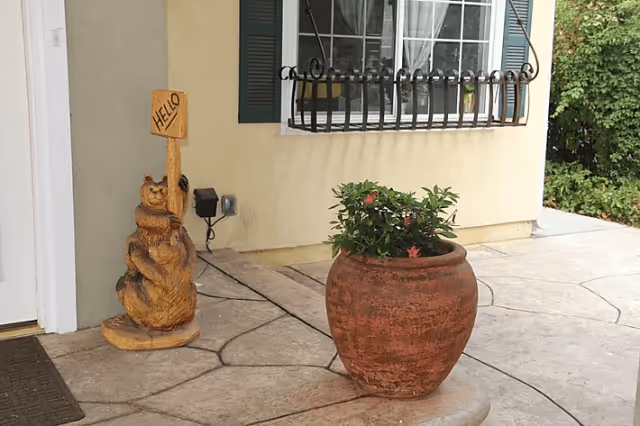 A terracotta pot with flowering plants sits on a tiled front porch beside a carved wooden bear holding a 'HELLO' sign under a window with decorative iron bars.