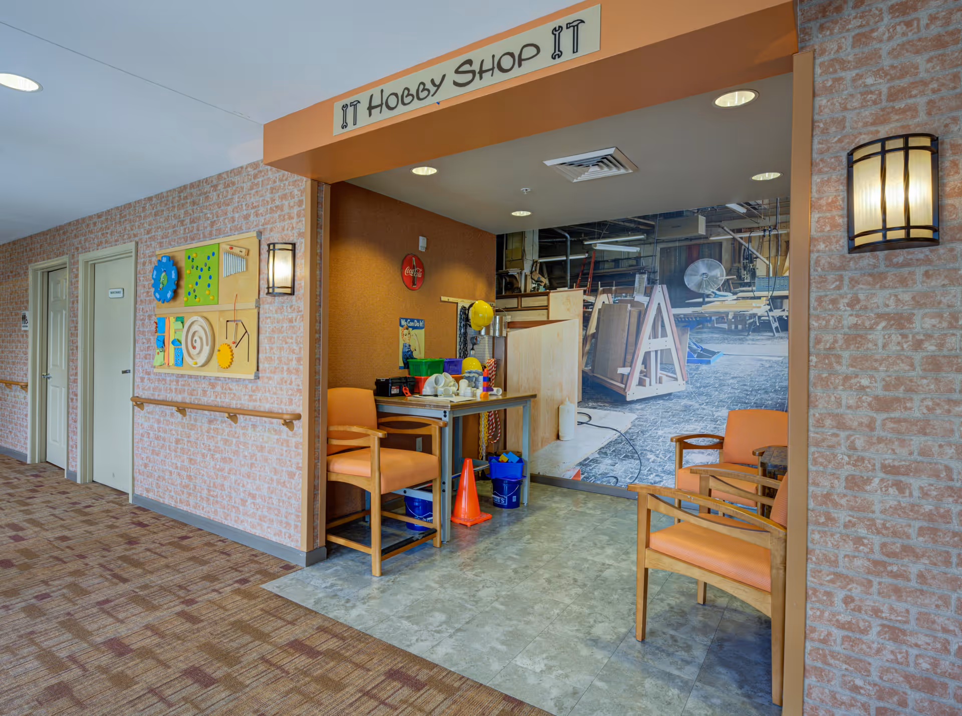 A hallway in a senior living facility with a small hobby shop area labeled 'IT Hobby Shop IT'. The hobby shop has two orange chairs, a table with various craft supplies, and a large wall mural depicting a workshop. The hallway has a carpeted floor, brick-patterned walls, and a handrail. There are two closed doors on the left side of the hallway and a colorful activity board mounted on the wall.