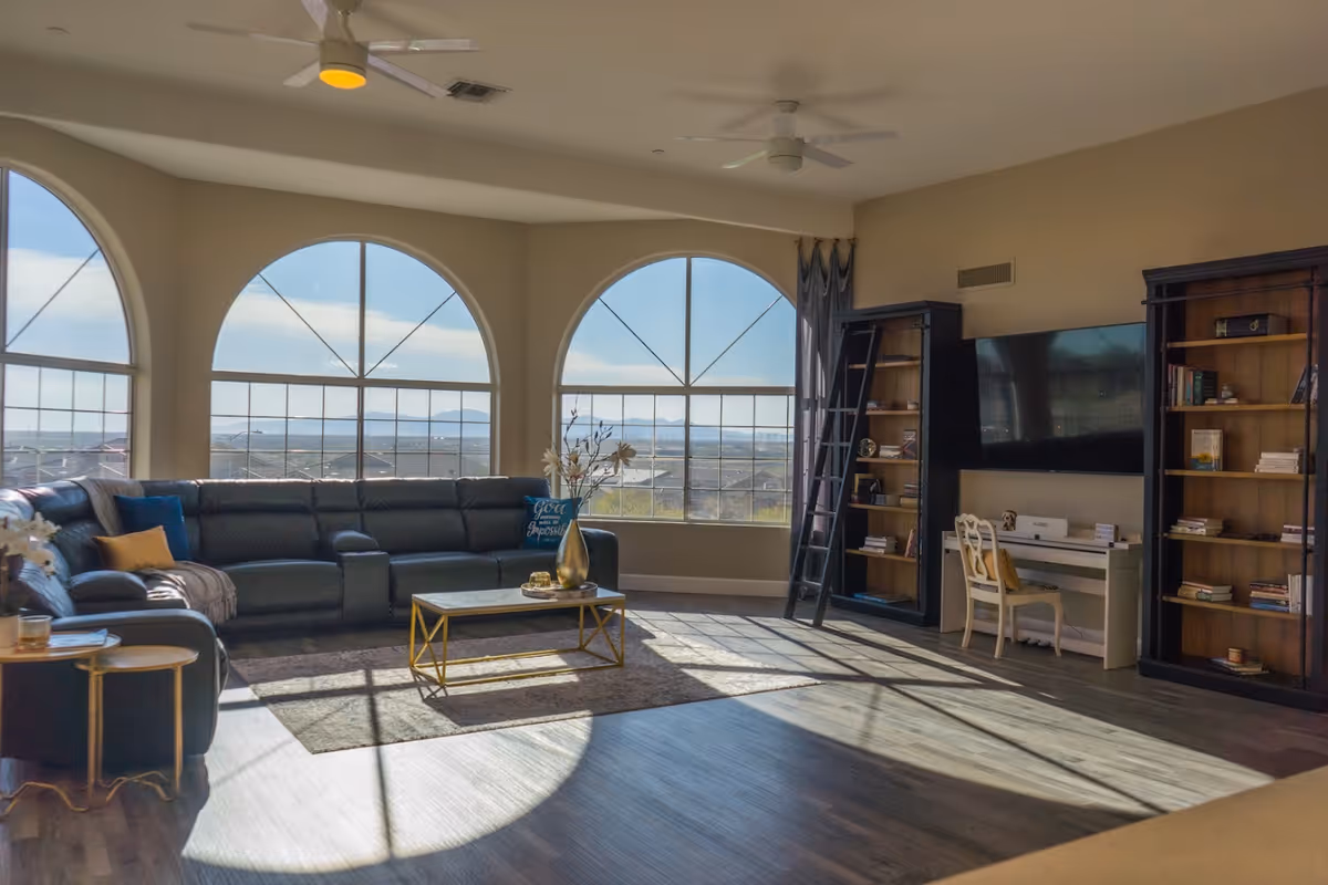 A spacious living room with large arched windows letting in natural light. The room features a dark sectional sofa with colorful pillows, a coffee table with a vase of flowers, two tall bookshelves with a rolling ladder, a wall-mounted flat-screen TV, and a small white desk with a chair. The floor is wooden, and ceiling fans are visible.