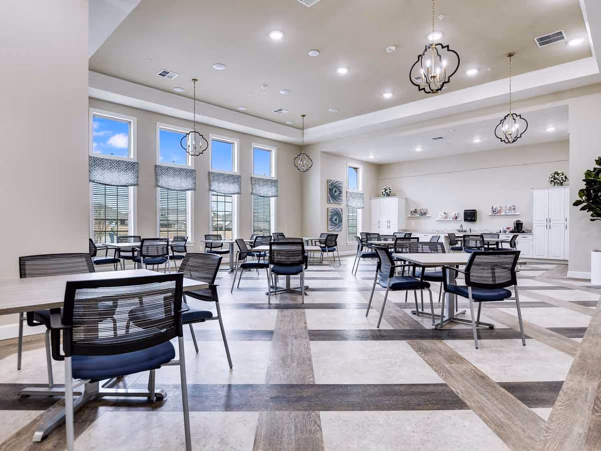 A spacious and bright dining room with multiple tables and chairs arranged neatly. Large windows with patterned valances allow natural light to fill the room. The ceiling features recessed lighting and decorative hanging light fixtures. The back wall has white cabinets and shelves with decorative items and a coffee station.