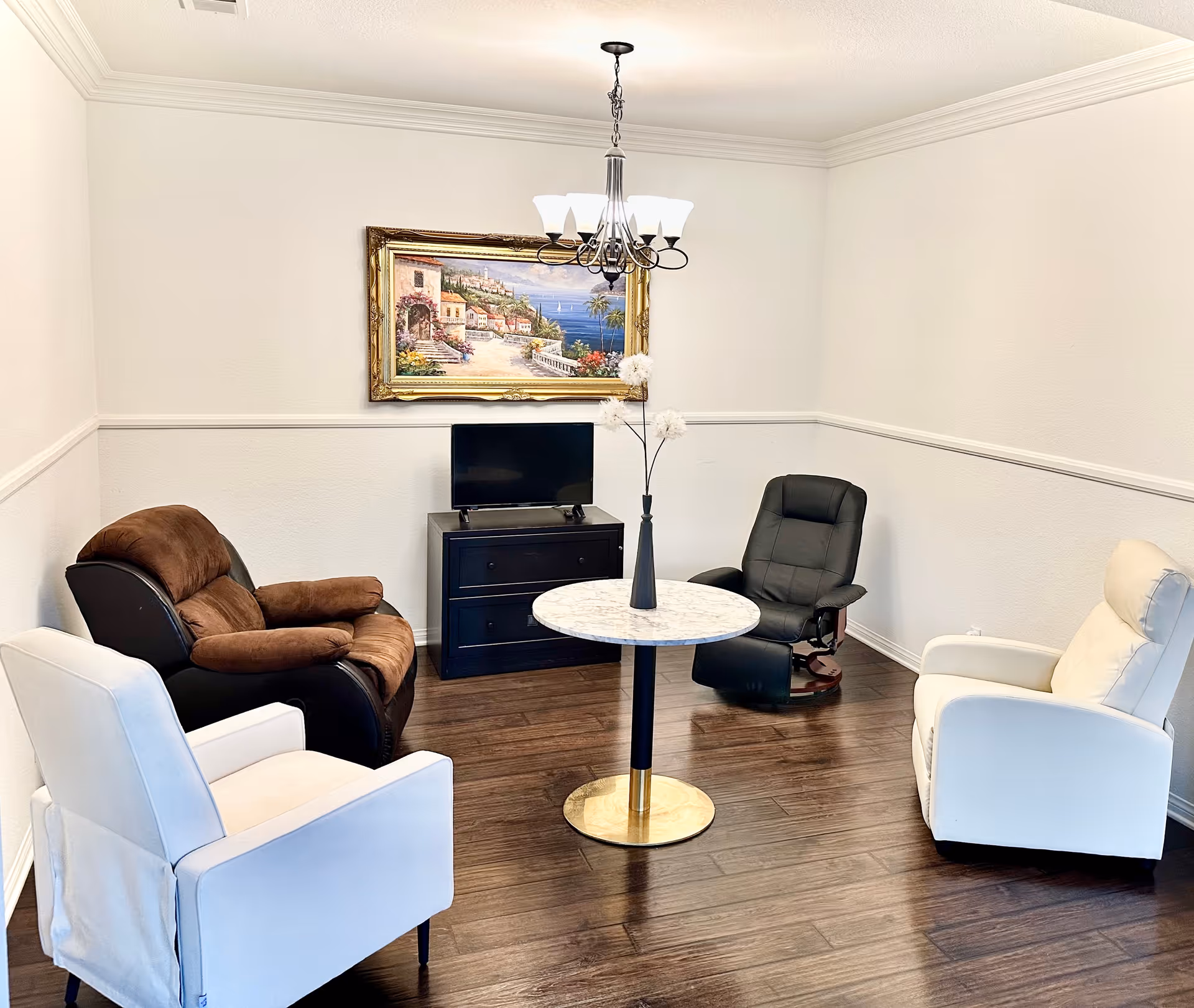 A small sitting area with four armchairs arranged around a round marble-top table with a vase holding white flowers. The room has white walls with a chair rail, dark wood flooring, a small black TV stand with a flat-screen TV, and a framed painting of a coastal village hanging above the TV. A chandelier with five lights hangs from the ceiling.