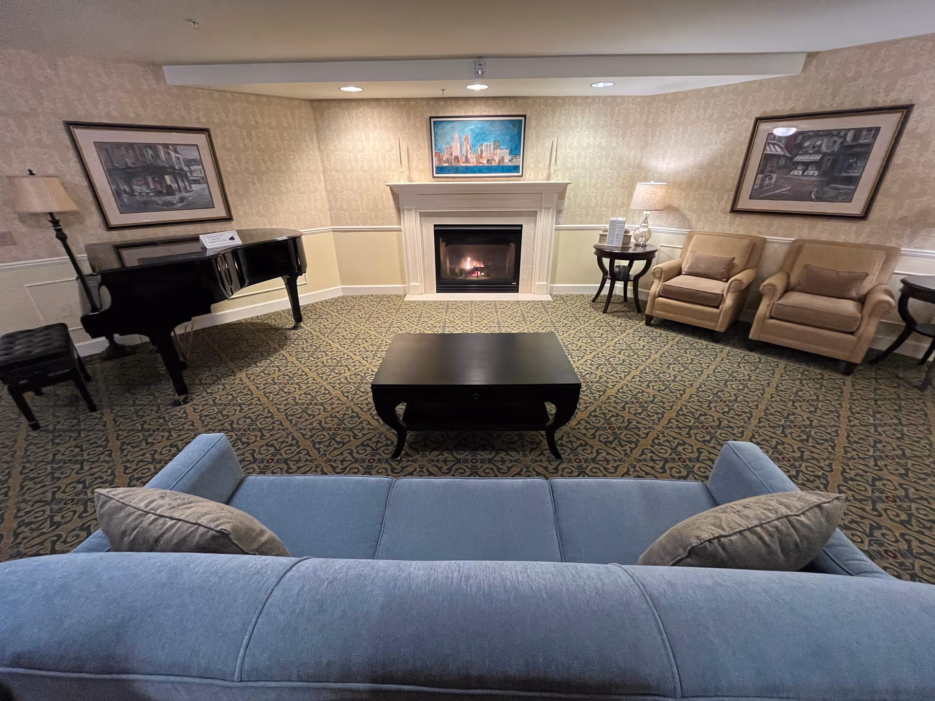 A cozy senior living community lounge with a blue sofa in the foreground, a black coffee table, two beige armchairs with pillows, a side table with a lamp, a grand piano with a bench, and a fireplace with a cityscape painting above it. The walls have patterned wallpaper and framed artwork.