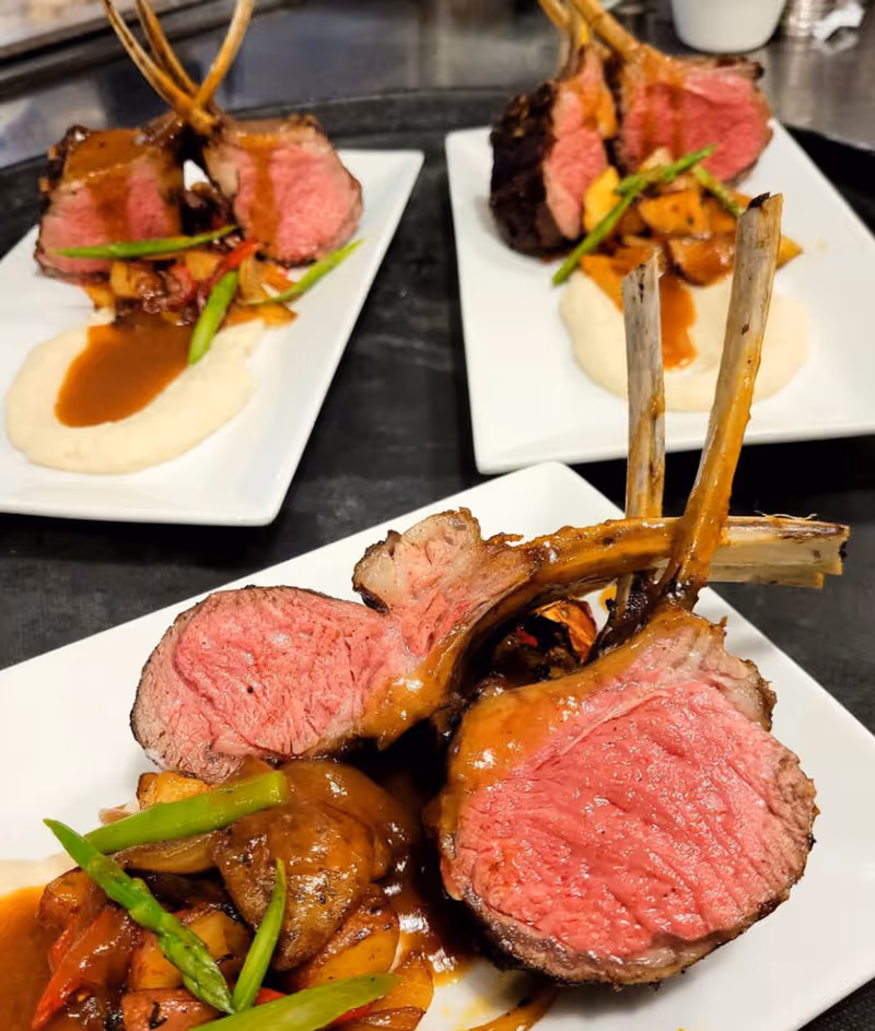 Three plates of gourmet lamb chops served with mashed potatoes, brown gravy, roasted vegetables, and asparagus spears on white rectangular plates.