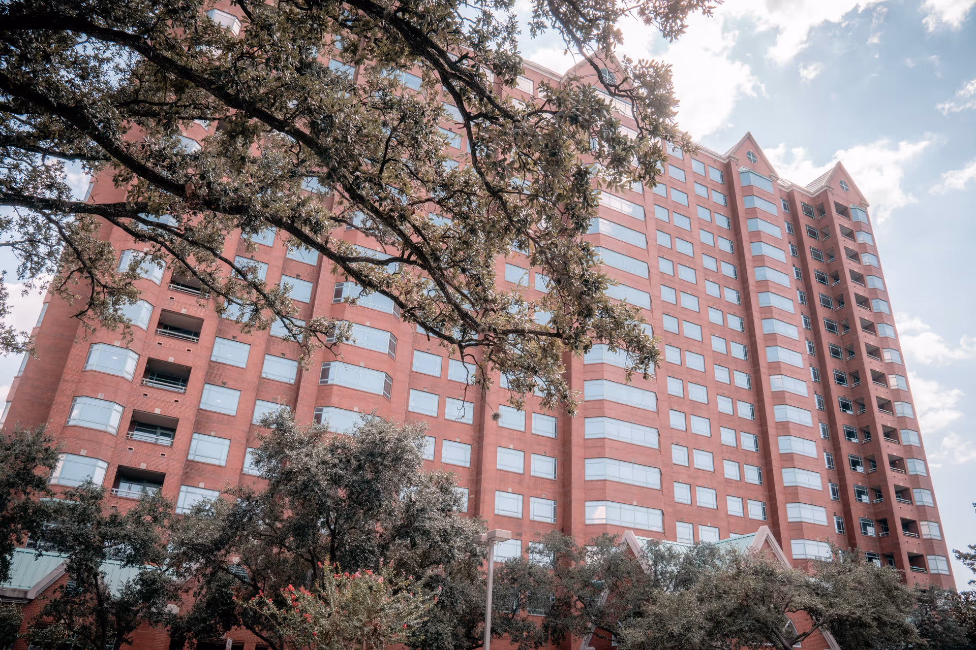 Exterior view of a tall red brick building with many windows, partially obscured by large trees with green leaves under a partly cloudy sky.