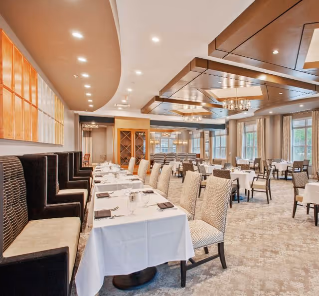 A spacious and elegant dining room with tables covered in white tablecloths, neatly set with glassware and napkins. The room features a mix of high-backed upholstered chairs and booth seating along one wall. Large windows with curtains allow natural light to fill the space, and decorative ceiling panels with chandeliers add a sophisticated touch.