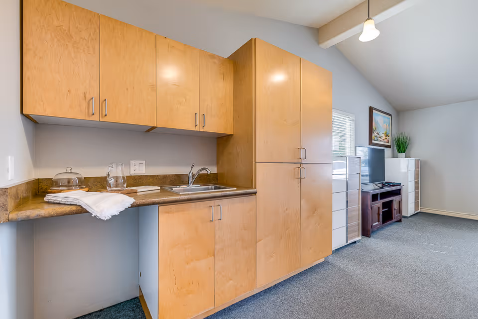 A small kitchenette with light wood cabinets, a sink and countertop opening into a carpeted living area with a TV and storage drawers.