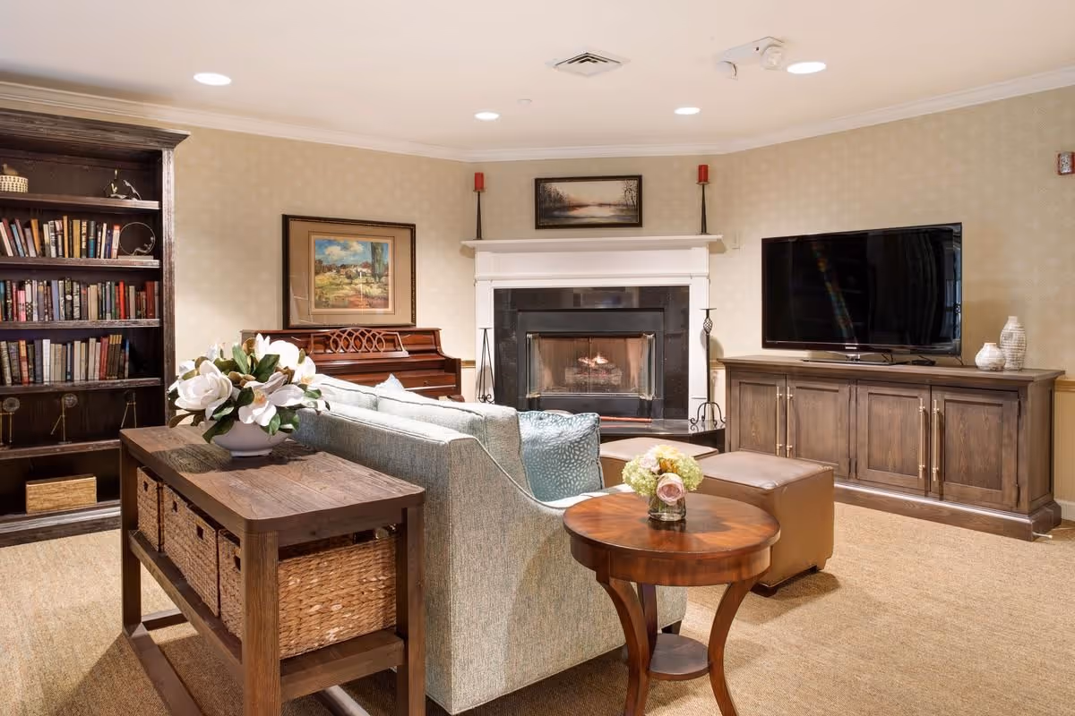 A cozy living room with a light blue sofa, a wooden side table with a small flower arrangement, a wooden console table with wicker baskets, a large flat-screen TV on a wooden cabinet, a fireplace with a black surround and white mantel, two red candles on tall holders, a piano, a bookshelf filled with books, and framed paintings on the walls.