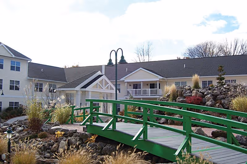 Outdoor view of a senior living facility named Legacy at Clover Blossom featuring a green wooden footbridge over a landscaped rocky garden area with ornamental grasses and plants, with the building and a covered entrance visible in the background under a partly cloudy sky.