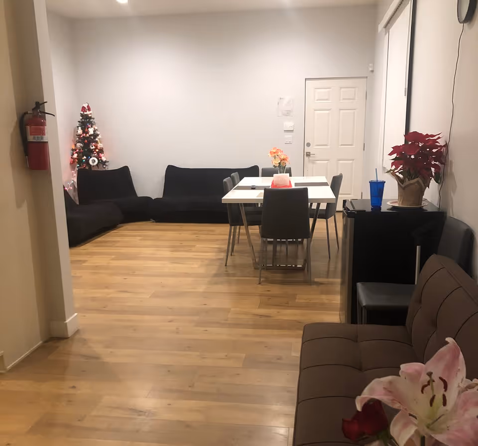 Interior view of a senior living facility room with wooden flooring, a black sectional sofa against the back wall, a white dining table with six chairs, a small decorated Christmas tree in the corner, a brown couch with flowers in the foreground, a small black refrigerator with a blue cup on top, and a fire extinguisher mounted on the left wall.