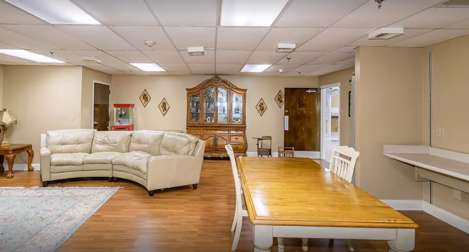 A senior living facility common area with a curved beige leather sofa, a wooden dining table with white chairs, a wooden china cabinet against the wall, a popcorn machine, and a small side table with a lamp. The room has wood flooring, beige walls, and a drop ceiling with fluorescent lights.