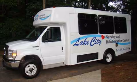 White bus labeled Lake City Nursing and Rehabilitation Center parked outdoors beside trees.