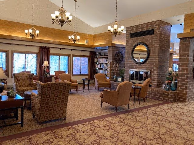 Cozy senior living common room with upholstered armchairs, a brick fireplace topped by a round mirror, bookshelves, chandeliers, and large windows.
