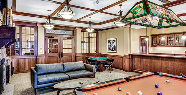 A cozy recreational room featuring a pool table with balls arranged on it, a black leather sofa with a wooden coffee table in front, a card table with four chairs in the background, and a bar area with hanging lights and glass cabinets. The room has wood paneling, beige walls, and multiple ceiling lights.