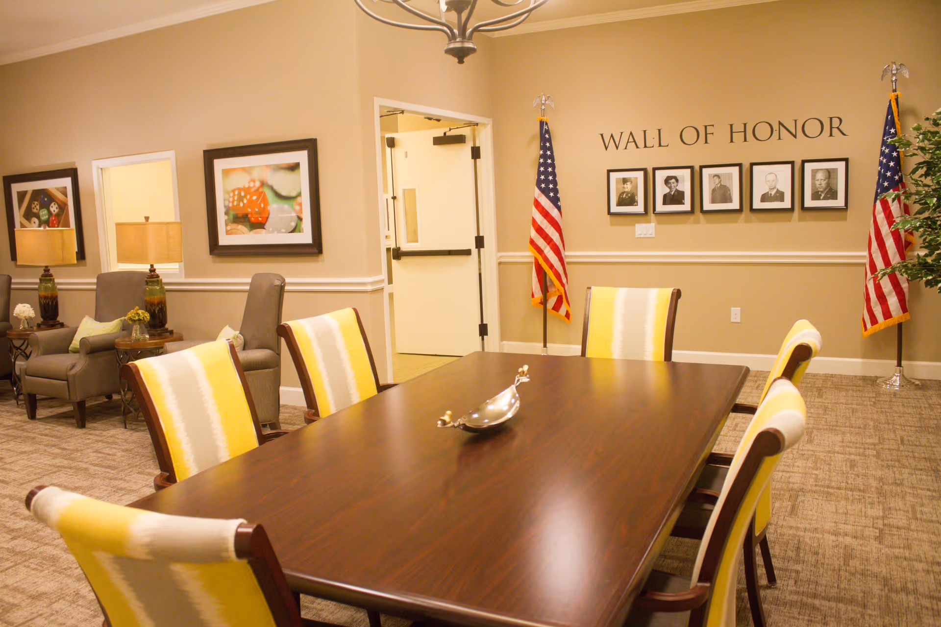 A well-lit room with a large wooden table surrounded by six yellow and white striped chairs. In the background, there are two American flags flanking a 'Wall of Honor' display with framed black and white photographs. To the left, there are two armchairs with side tables and lamps, and framed artwork on the wall.