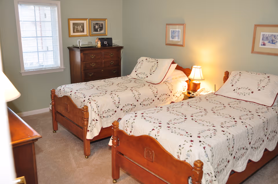 A bedroom with two wooden twin beds covered with matching white quilts featuring a floral pattern. Between the beds is a nightstand with a lamp and a telephone. There is a wooden dresser against the wall with framed pictures above it and a window with blinds on the left side.