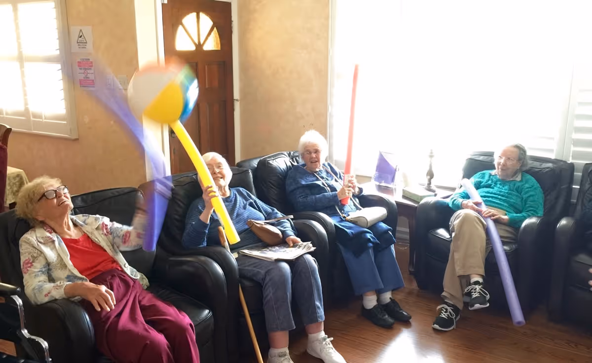 Four elderly women sitting in black leather armchairs in a living room with wooden floors and a wooden door. They are playing with colorful foam pool noodles and a beach ball, appearing engaged and cheerful. The room has beige walls and windows with white shutters letting in natural light.